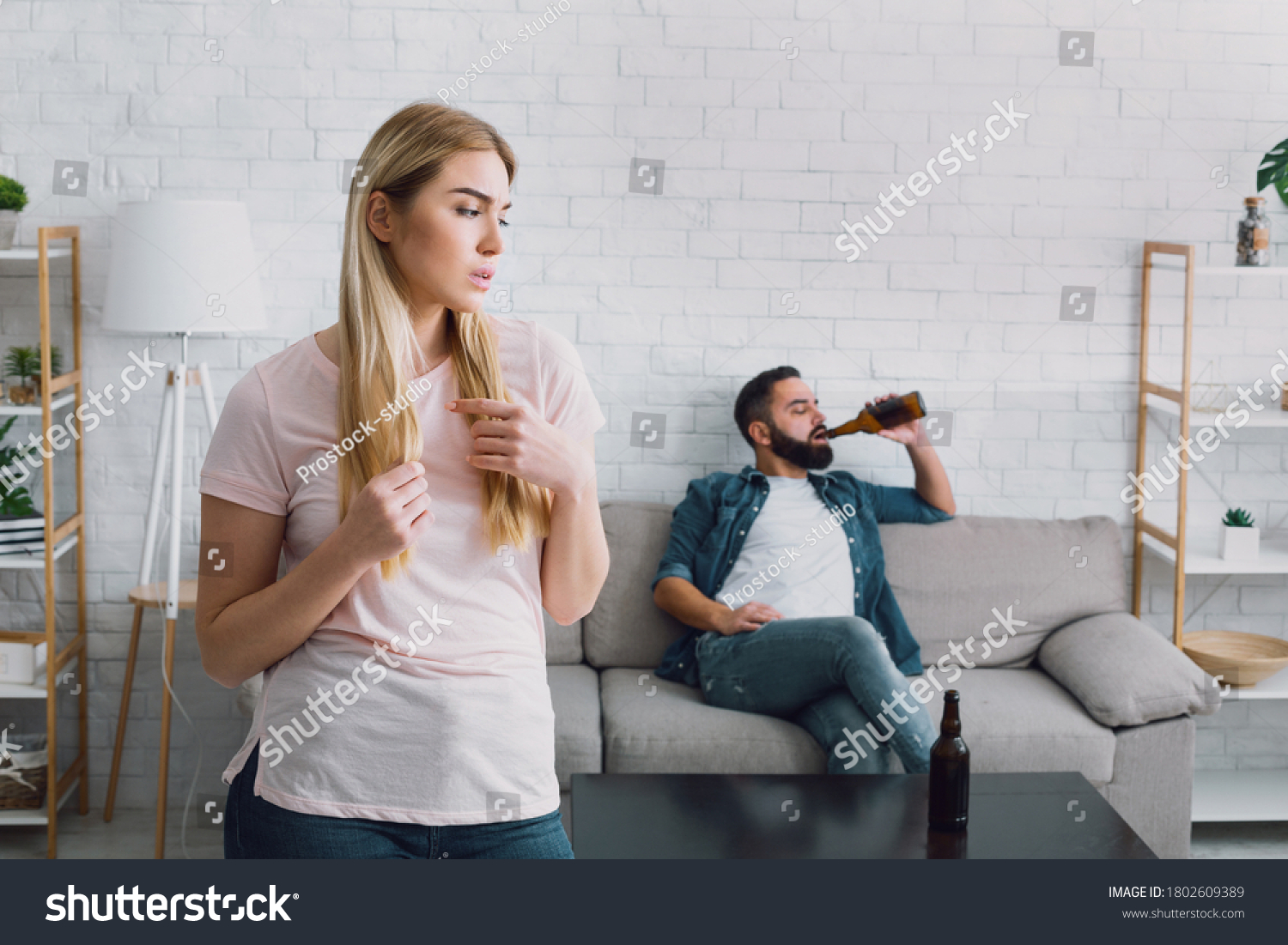 Latent alcoholism. Sad wife sees her husband drinking a lot of beer, man with beard sits on couch, near table with empty bottles #1802609389