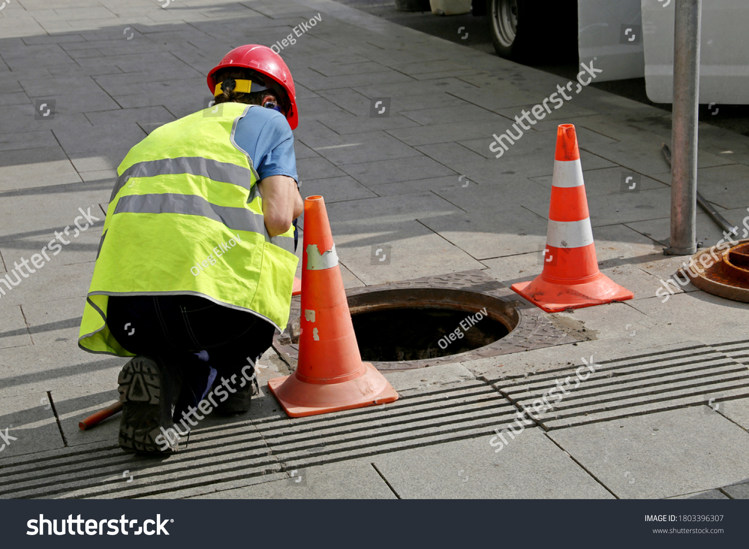 Worker over the open sewer hatch on a street near the traffic cones. Concept of repair of sewage, underground utilities, water supply system, cable laying, water pipe accident #1803396307