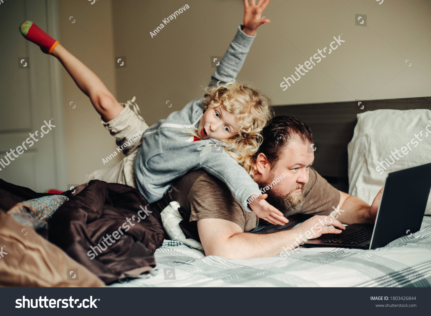 Work from home with kids children. Father working on laptop in bedroom with child daughter on his back. Funny candid family moment. New normal during coronavirus self-isolation quarantine. #1803426844
