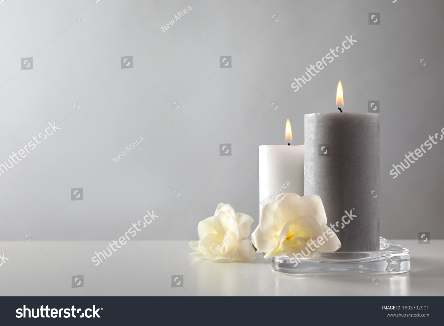Wax candles and flowers in glass holder on table against light background. Space for text #1803792901