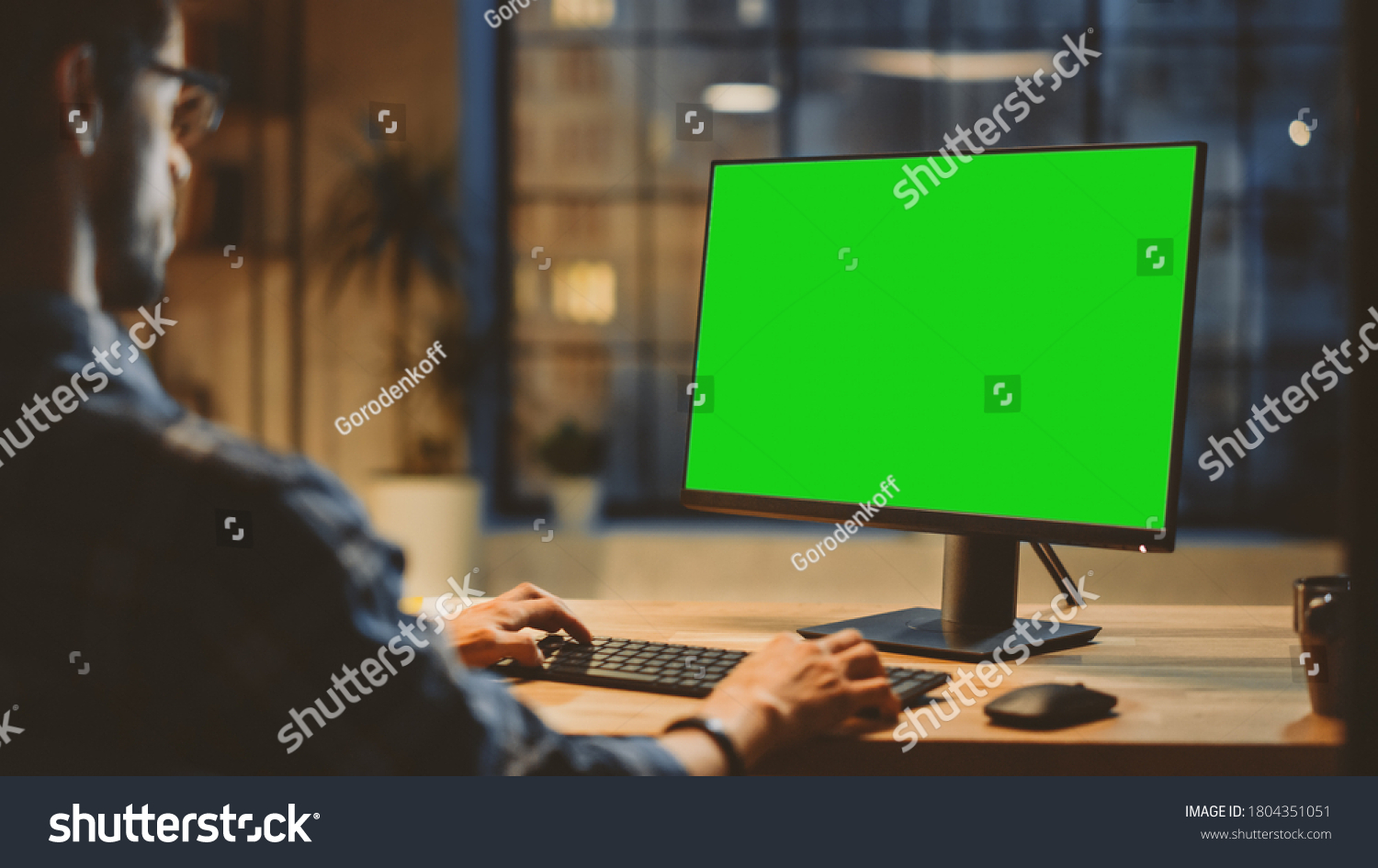 Over the Shoulder: Creative Young Man Sitting at His Desk Using Desktop Computer with Mock-up Green Screen. Evening in the Stylish Office Studio with City Window View #1804351051