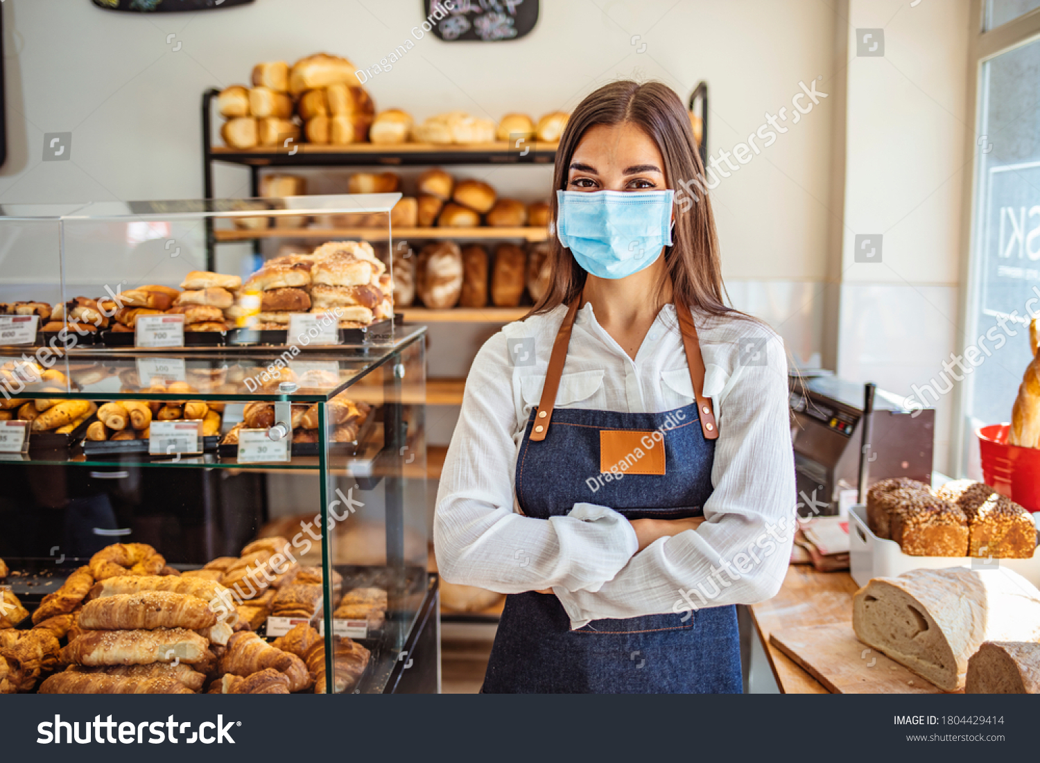 Woman working at a bakery wearing a facemask to avoid the coronavirus. COVID-19 lifestyle concepts. Day in the life of owners of bakery shop with the protocol against the Covid-19 in place.  #1804429414