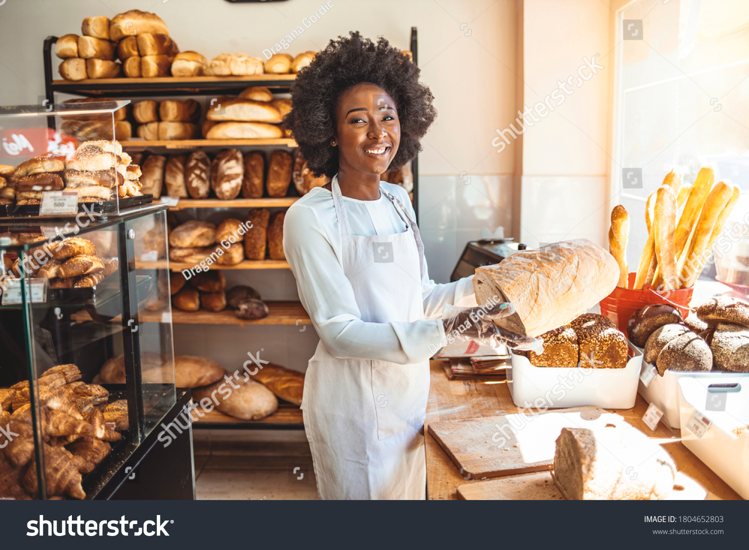 Smiling baker woman standing with fresh bread at bakery. Happy african woman standing in her bake shop and looking at camera. Satisfied baker with breads in background. Beautiful  woman at bakery shop #1804652803