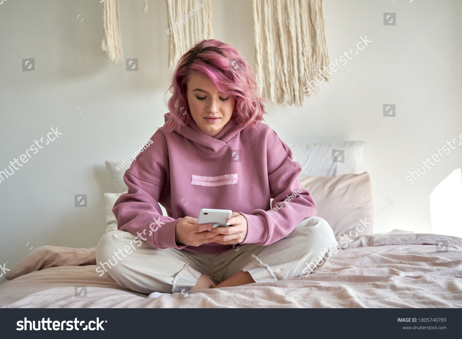 Hipster woman with pink hair holding smart phone chatting in social media, watching video content, texting messages, playing games, using mobile apps, surfing internet sit in bed at home. Vertical #1805740789