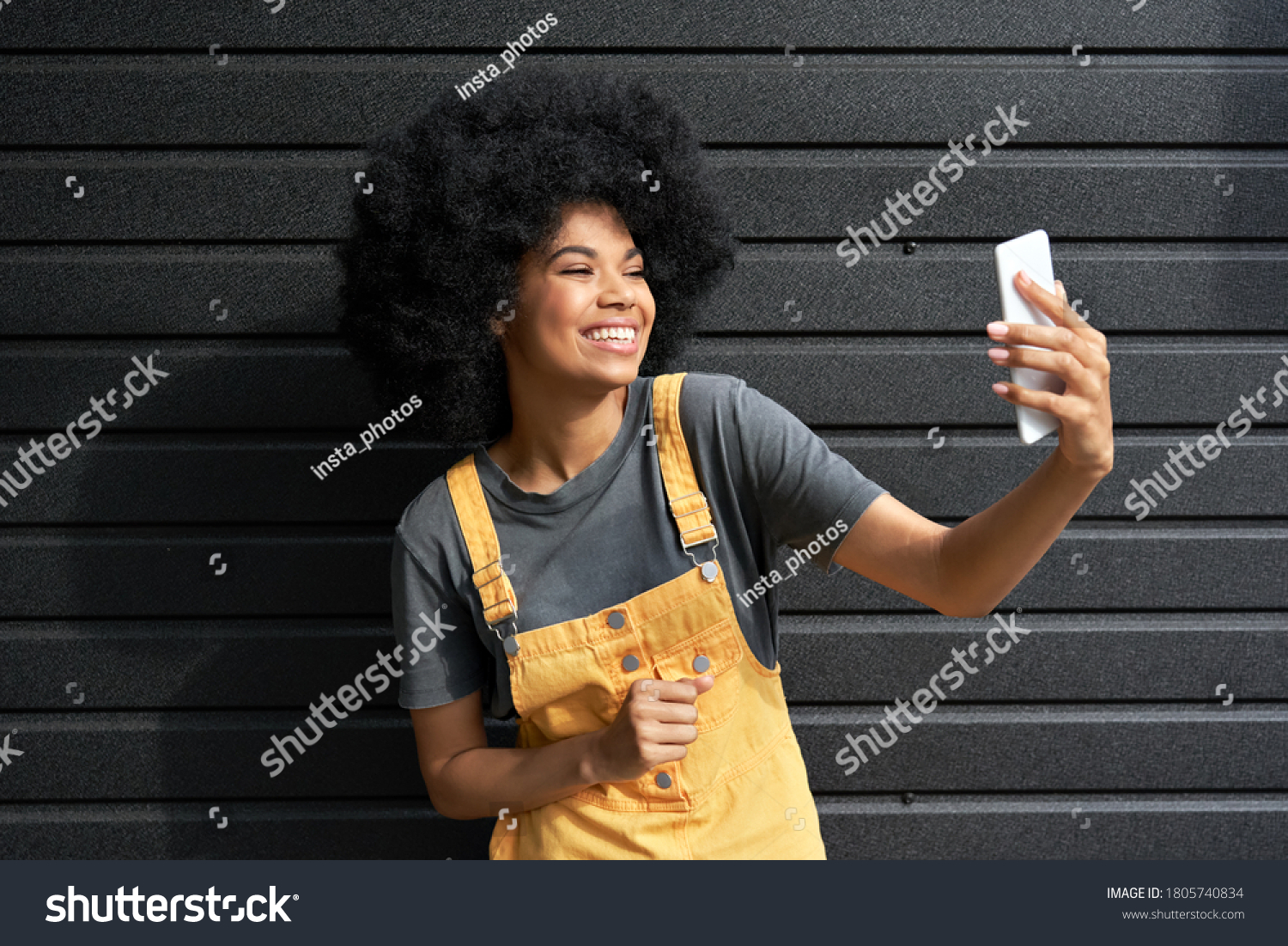 Happy African American hipster woman blogger with Afro hair laughing, holding phone, recording vlog, shooting social media stories or video calling in mobile app standing on black background. #1805740834