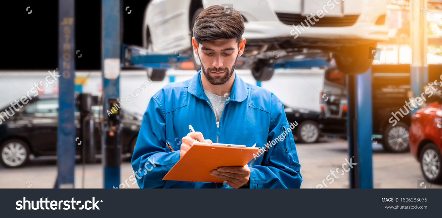 Mechanic in blue work wear uniform checks the vehicle maintenance checklist with blur lifted car in the background. Automobile repairing service, Professional occupation. #1806288076