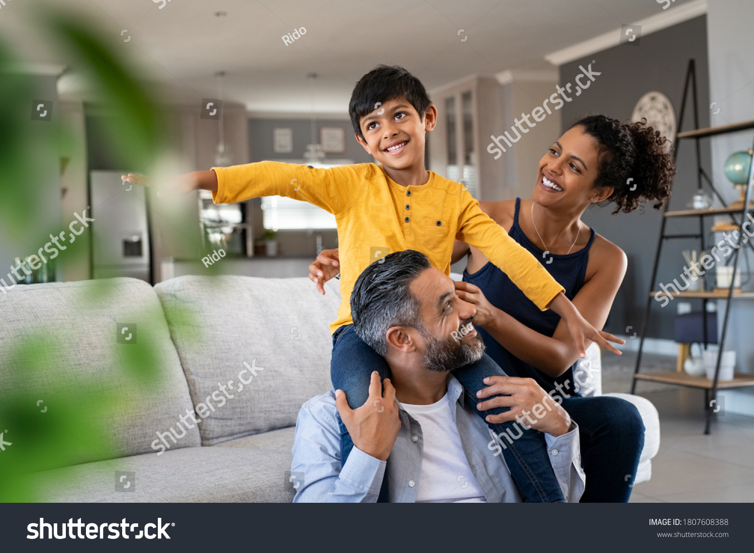 Cheerful indian son sitting on father shoulder playing at home with african mother. Playful little boy enjoying spending time with parents at home. Flying child enjoying playing with his ethnic family #1807608388