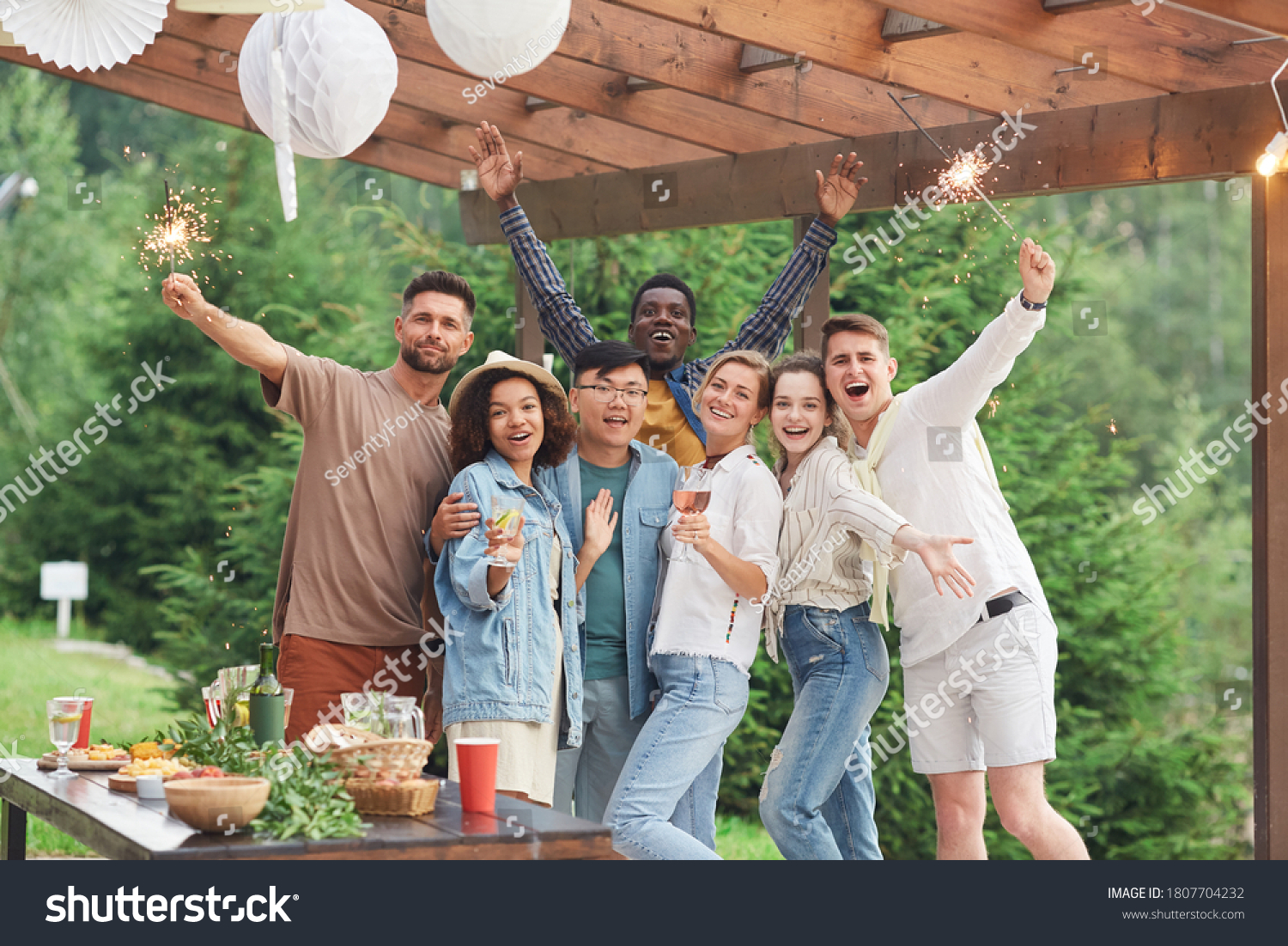 Multi-ethnic group of happy friends holding sparklers and looking at camera while enjoying Summer party outdoors, copy space #1807704232