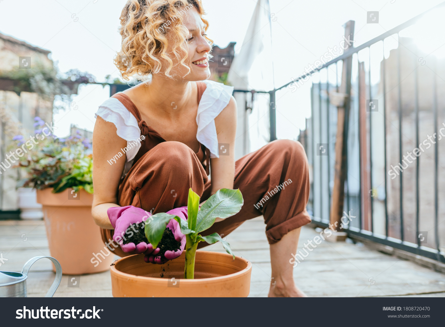 Pleasant active resting terrace. Blond romantic female gardener in overalls enjoying hobbie, creating garden at balcony. Plants cultivation and nursery, garden maintenance. #1808720470