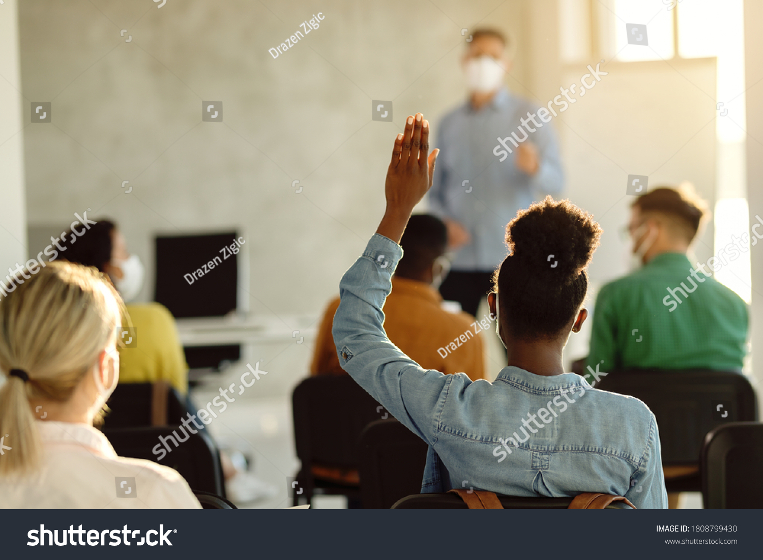 Back view of black female student raising arm to ask a question during a lecture in the classroom.  #1808799430