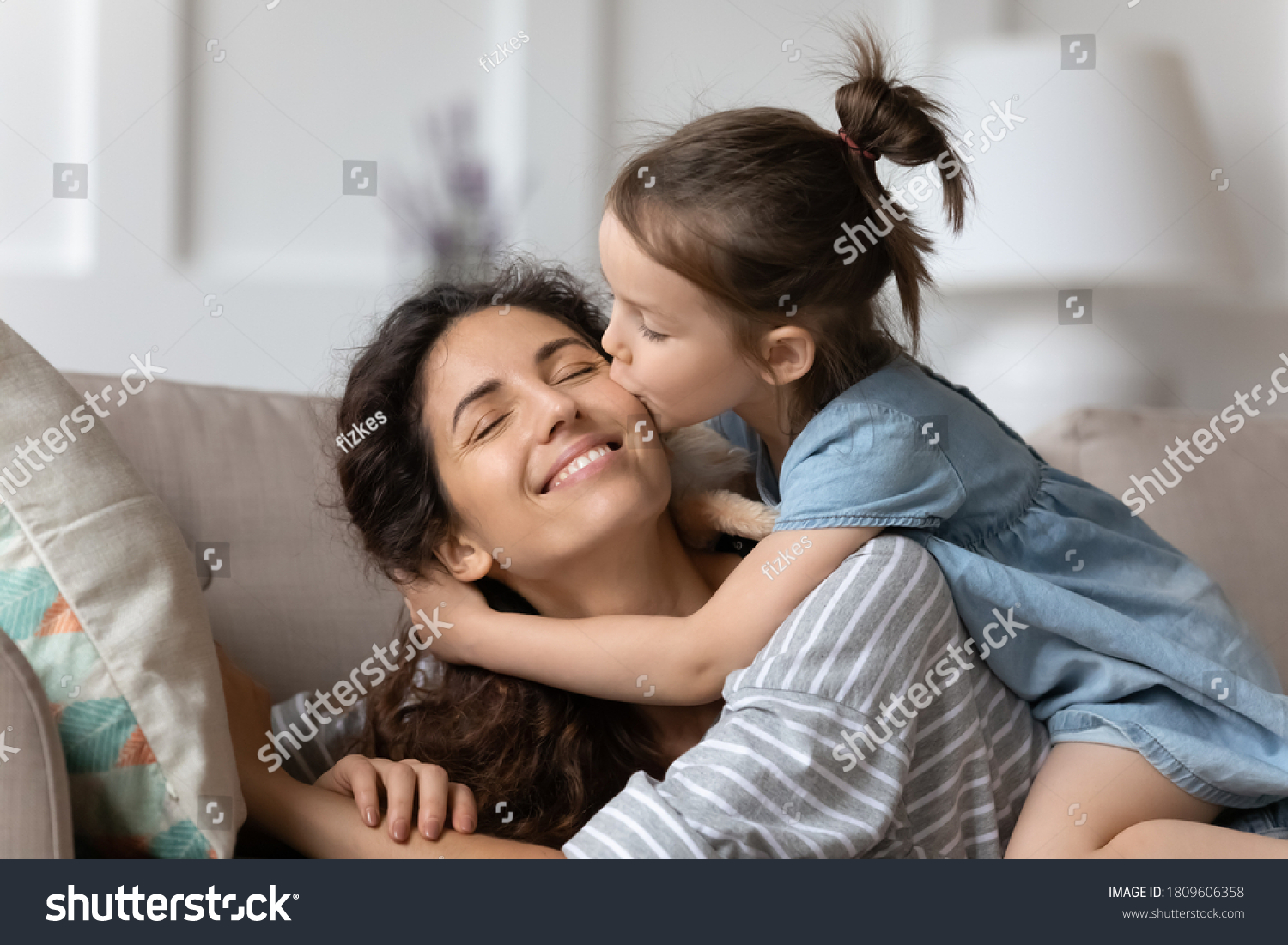 Little daughter gently cuddles kiss mother on cheek showing love and express caress resting on couch at home. Happy family, pleasure be mommy, mother day congratulations, sweet moment together concept #1809606358