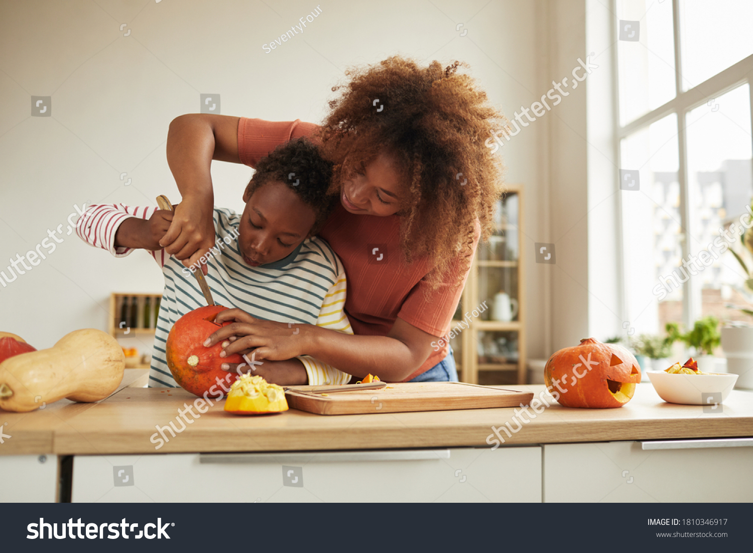 Stylish African American woman spending time with her son standing at table carving pumpkin for Halloween with kitchen knife together #1810346917