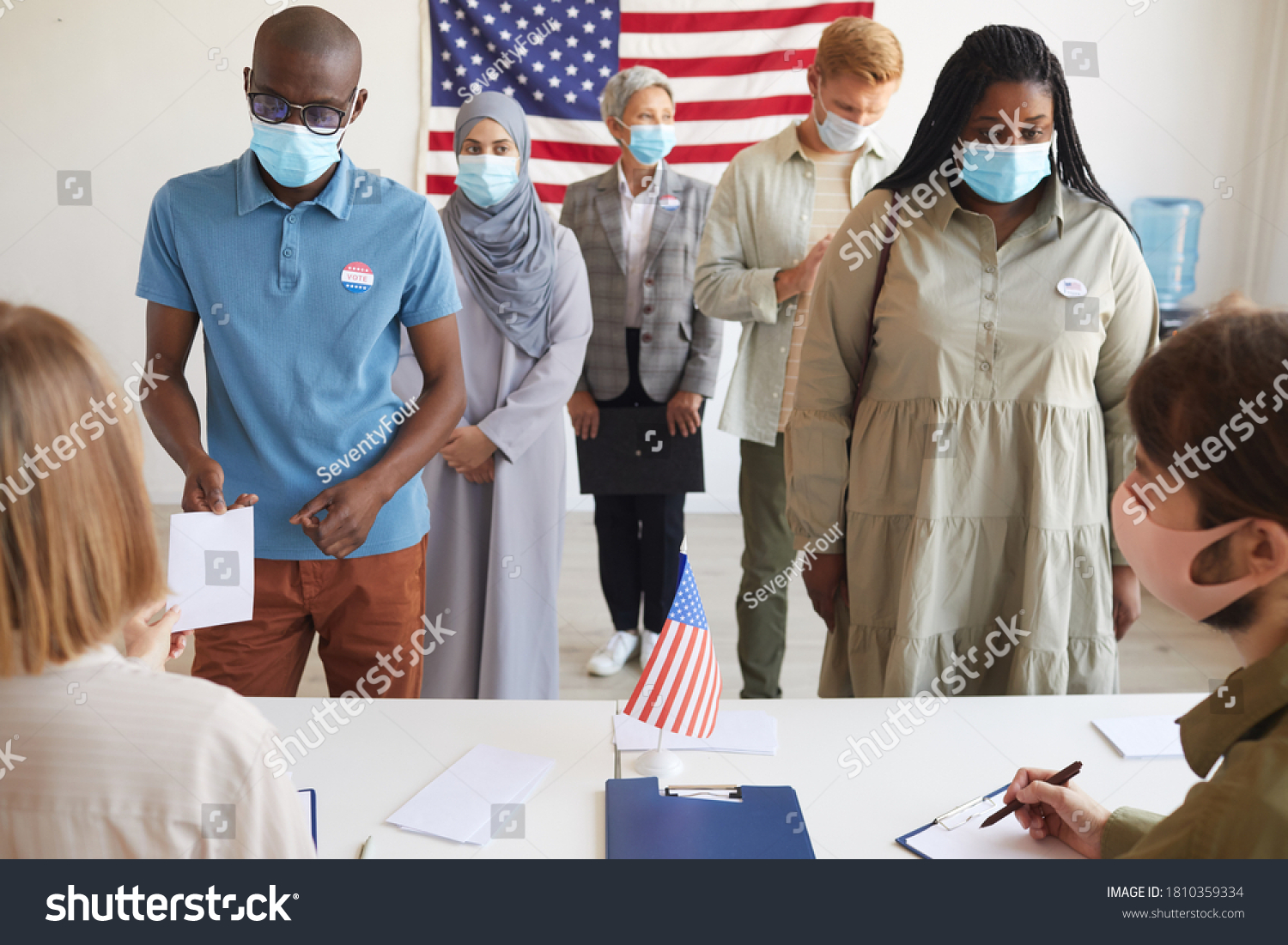 Front view at multi-ethnic group of people standing in row and wearing masks at polling station on election day, focus on two African-American people registering for voting #1810359334