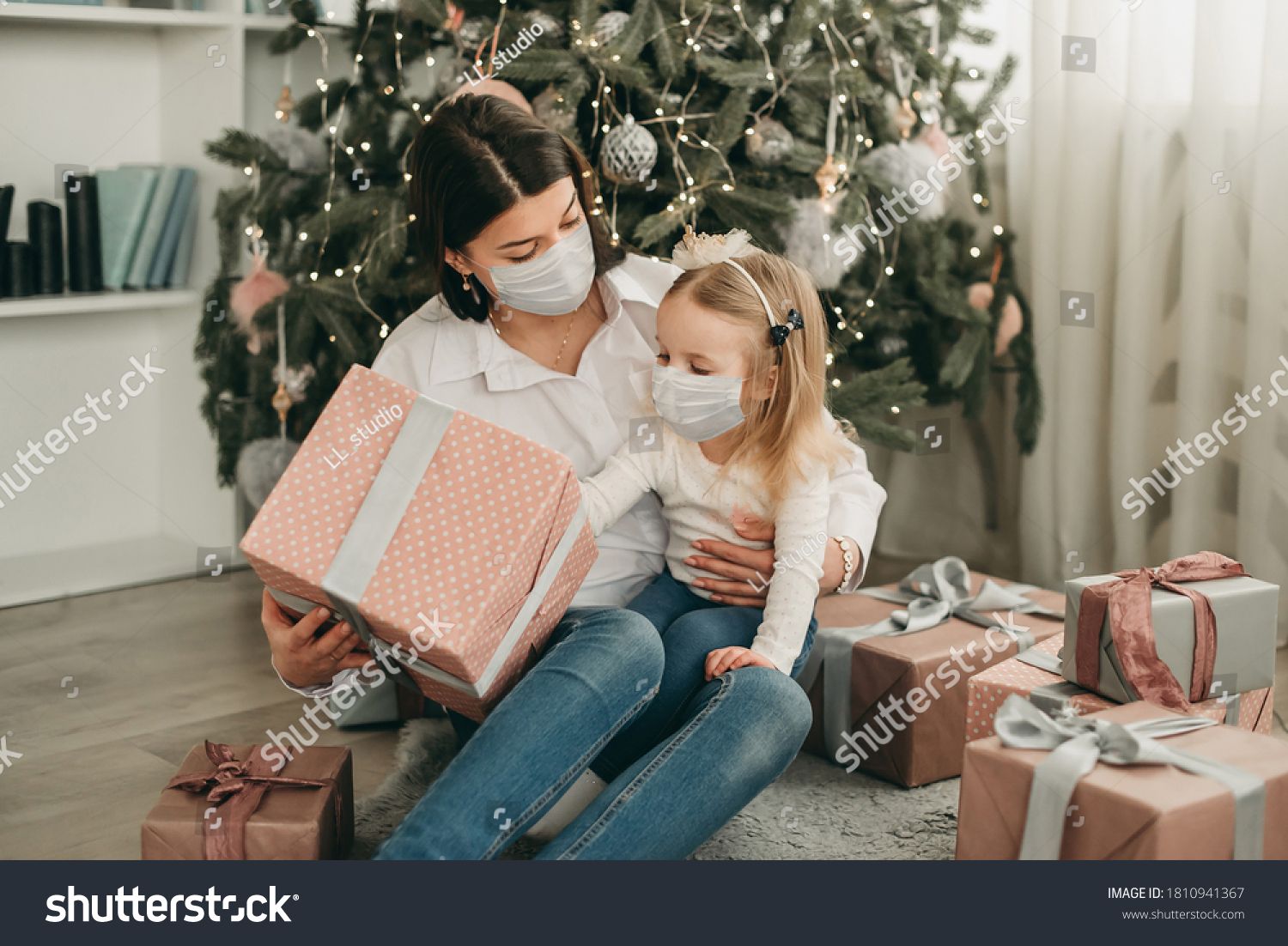 Beautiful mother and daughter in medical masks have fun at home near the Christmas tree in a white interior. Family happiness, holiday, joy, vacation, games with a woman. New Year's preparations. Cele #1810941367