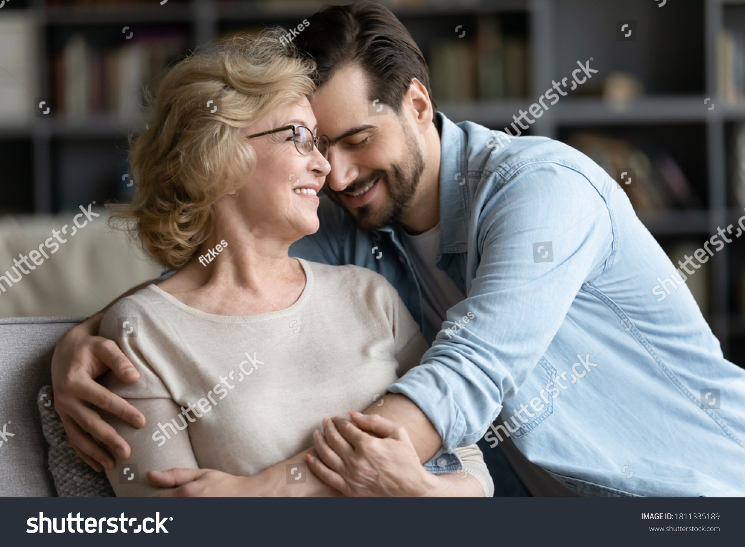 Smiling mature woman and adult son cuddling, enjoying tender moment, sitting on cozy couch in living room, happy beautiful elderly mother wearing glasses and young man hugging, two generations #1811335189
