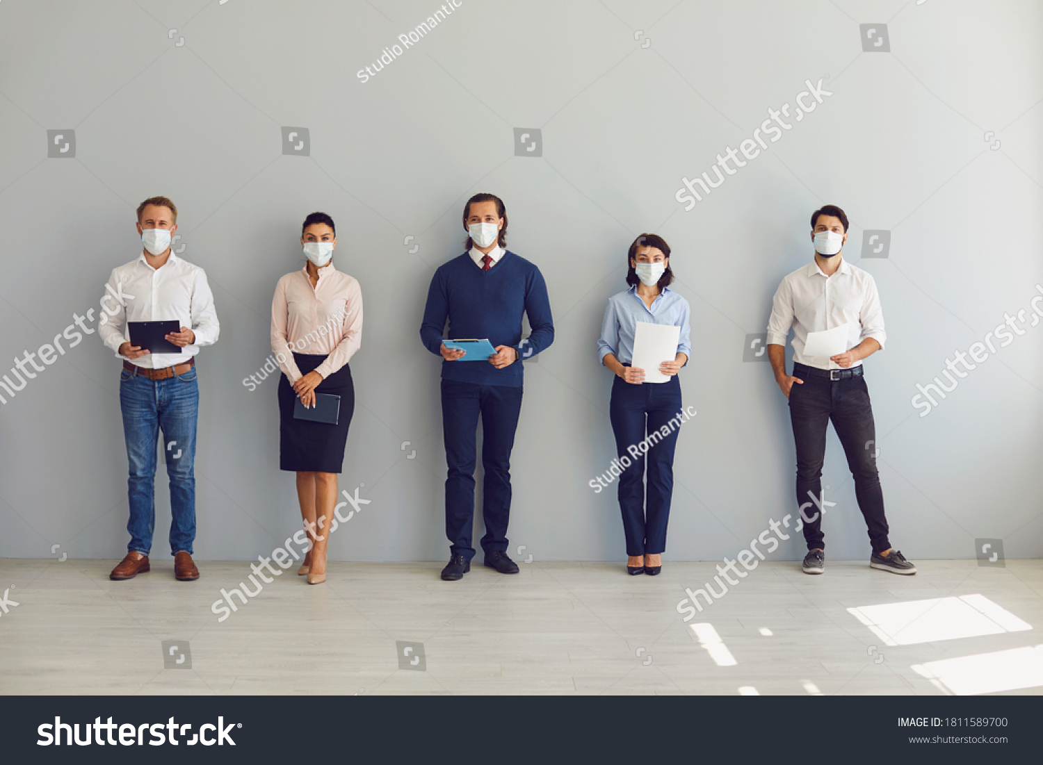 Social Distancing at Work concept. Office workers or job candidates in protective face masks standing in corridor. Applicants waiting for interview keeping safe distance to prevent spread of Covid 19 #1811589700
