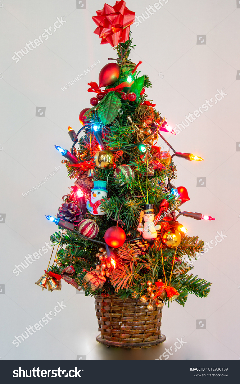 Green Christmas tree with lights decorated with typical figures of the date, with red balls, with mini gifts, and a red bow at the top of the tree with a white background. Vertical photography. #1812936109