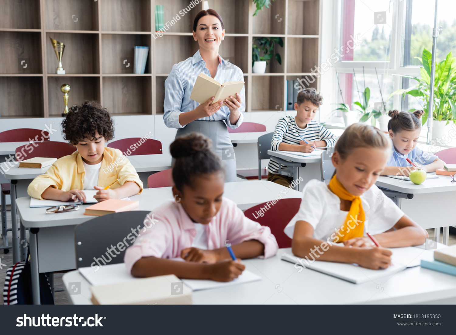 teacher holding book and looking at camera while multicultural pupils writing dictation in classroom #1813185850