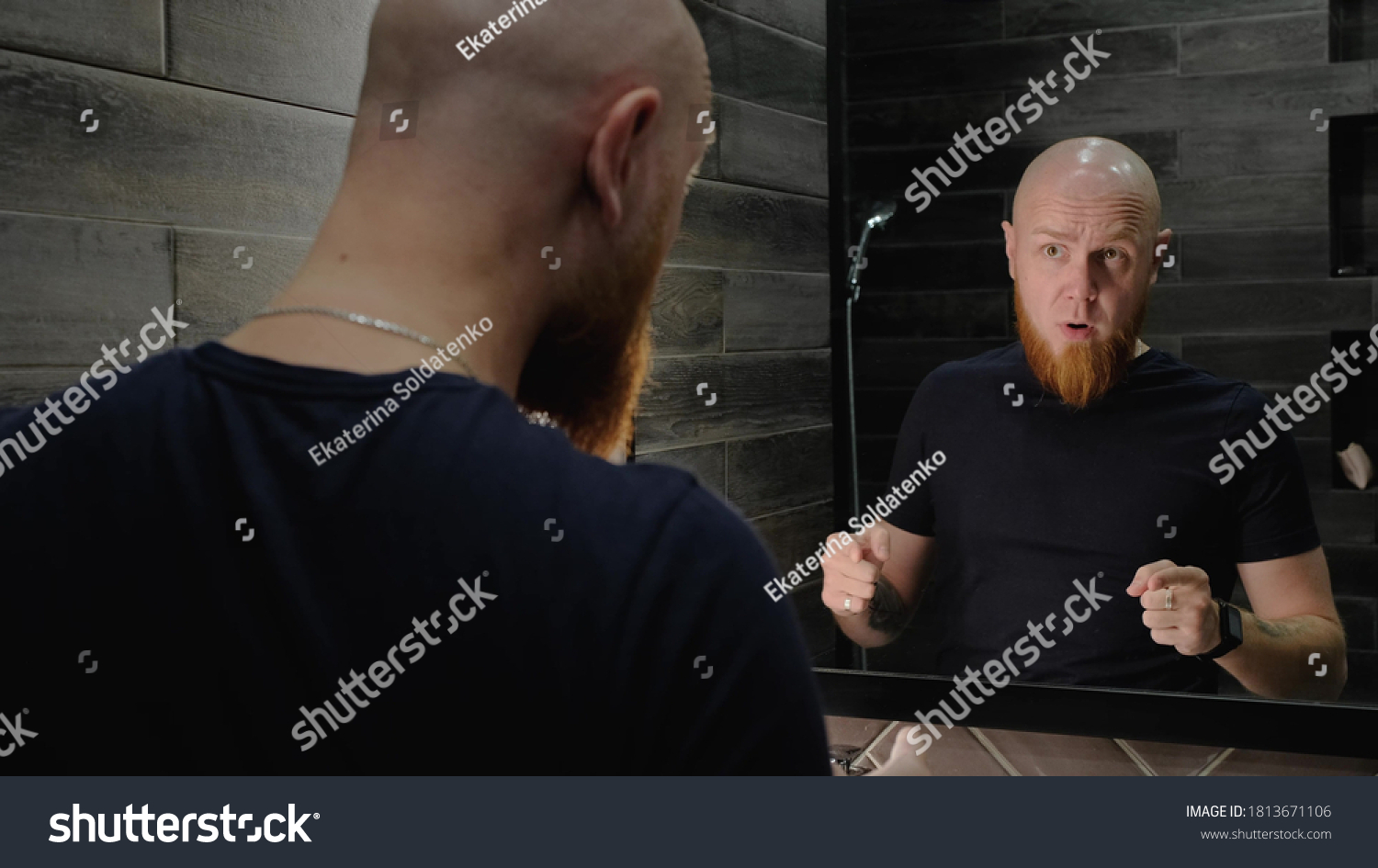 A young, Caucasian, charismatic, bald guy with a red beard invigorates himself in front of the mirror, talking to his reflection in a modern bathroom. Morning mood for the day. Recharge with energy. #1813671106