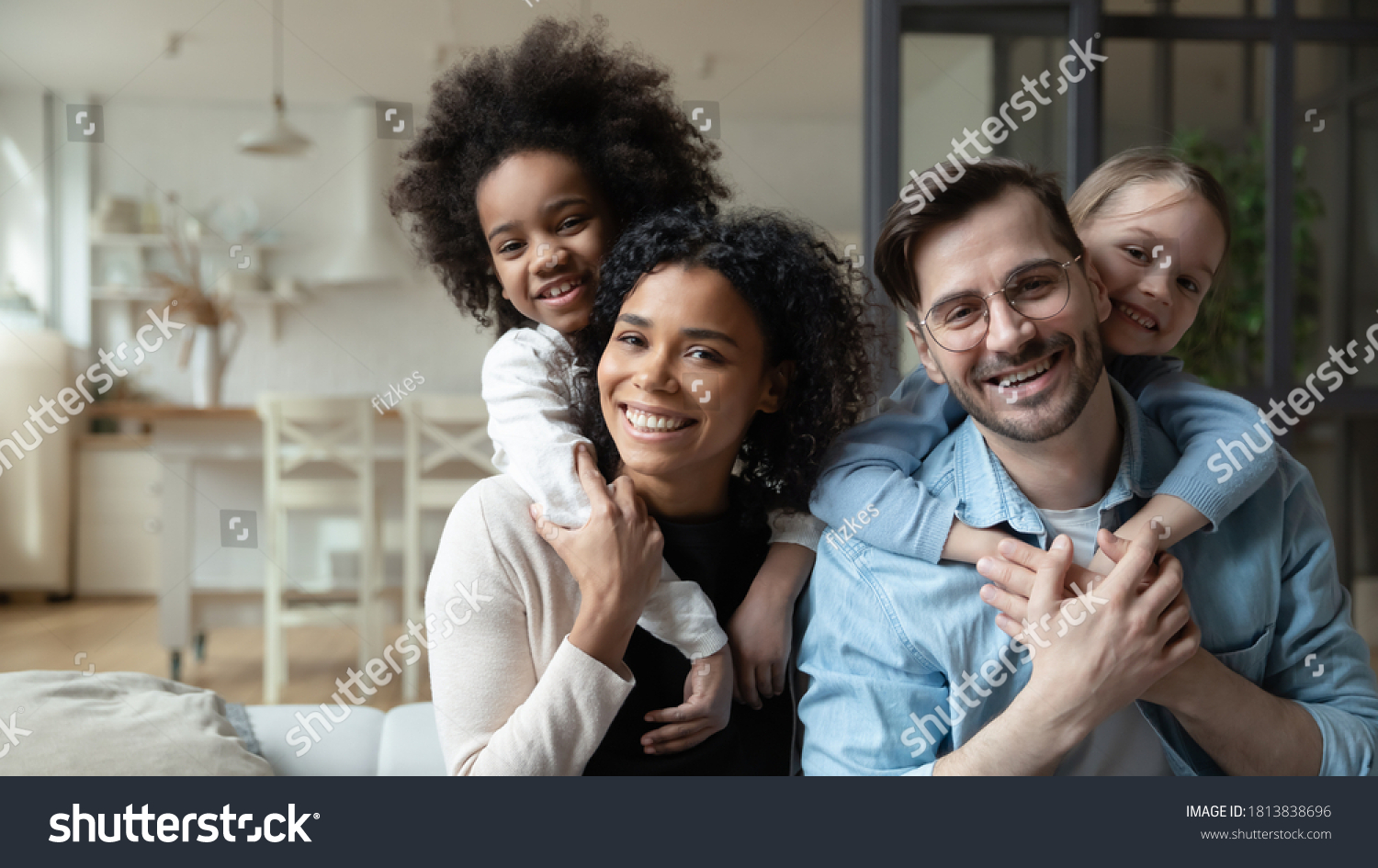 Portrait of happy multiracial couple enjoying sweet family moment with adorable little mixed raced daughters at home. Smiling cute small stepsisters cuddling cheerful parents, looking at camera. #1813838696