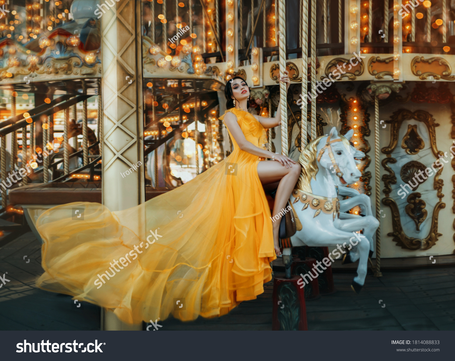 Young beautiful stylish woman sits astride a toy horse, rides a carousel. Long bright yellow dress fluttering in motion #1814088833