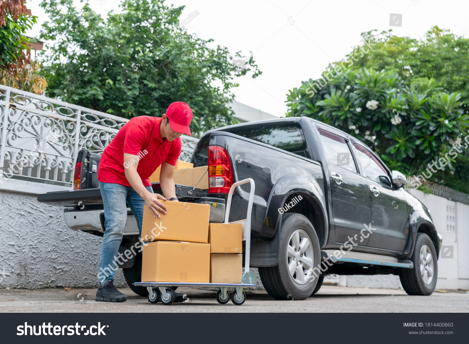 Delivery men in red uniform unloading cardboard boxes from pickup truck. Courier man sending the parcel or package to the customer on a business day. Online shopping and transport logistics concept. #1814400860