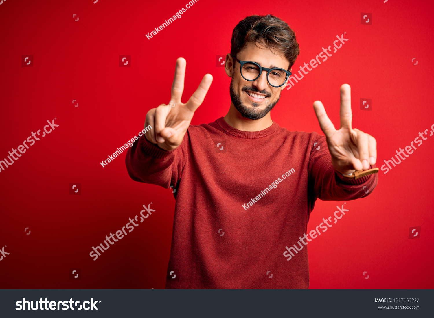 Young handsome man with beard wearing glasses and sweater standing over red background smiling with tongue out showing fingers of both hands doing victory sign. Number two. #1817153222