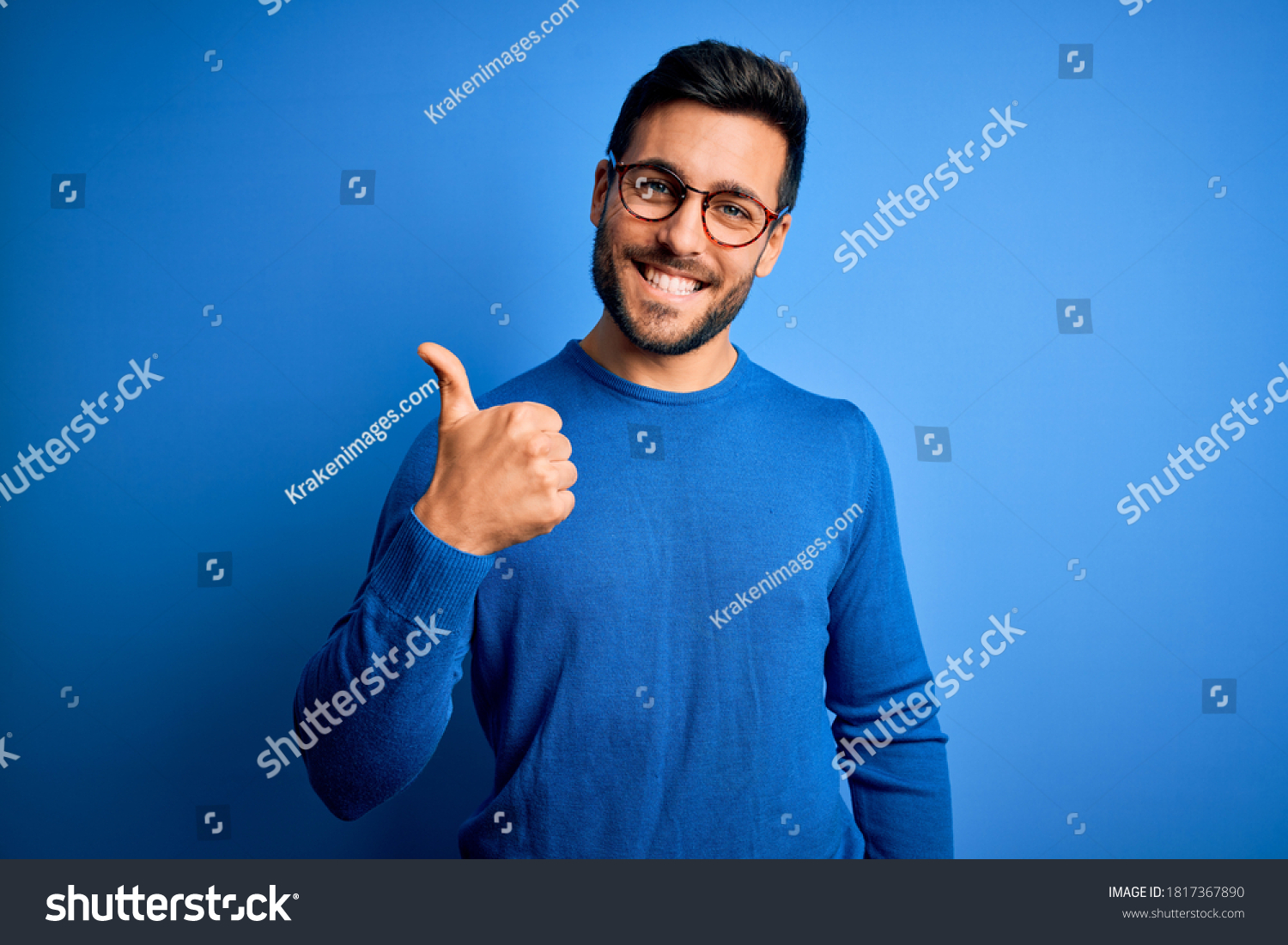 Young handsome man with beard wearing casual sweater and glasses over blue background doing happy thumbs up gesture with hand. Approving expression looking at the camera showing success. #1817367890
