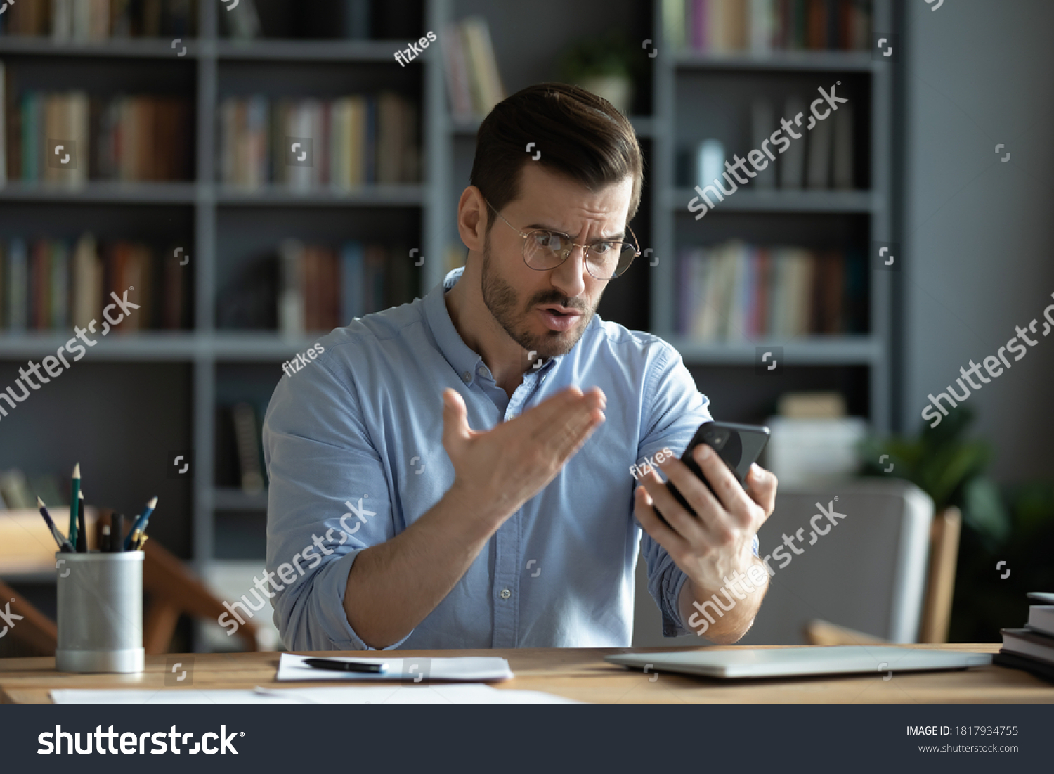 Mad millennial Caucasian male sit at desk look at cellphone shocked frustrated by gadget operational problems. Angry man stressed by message or error on smartphone, confused by slow internet. #1817934755