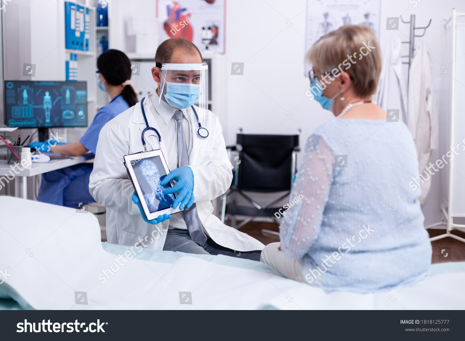 Doctor using tablet pc during examination of x-ray for older patient in hospital room and wearing face mask against coronavirus pandemic. Medical examination for infections, disease and diagnosis. #1818125777