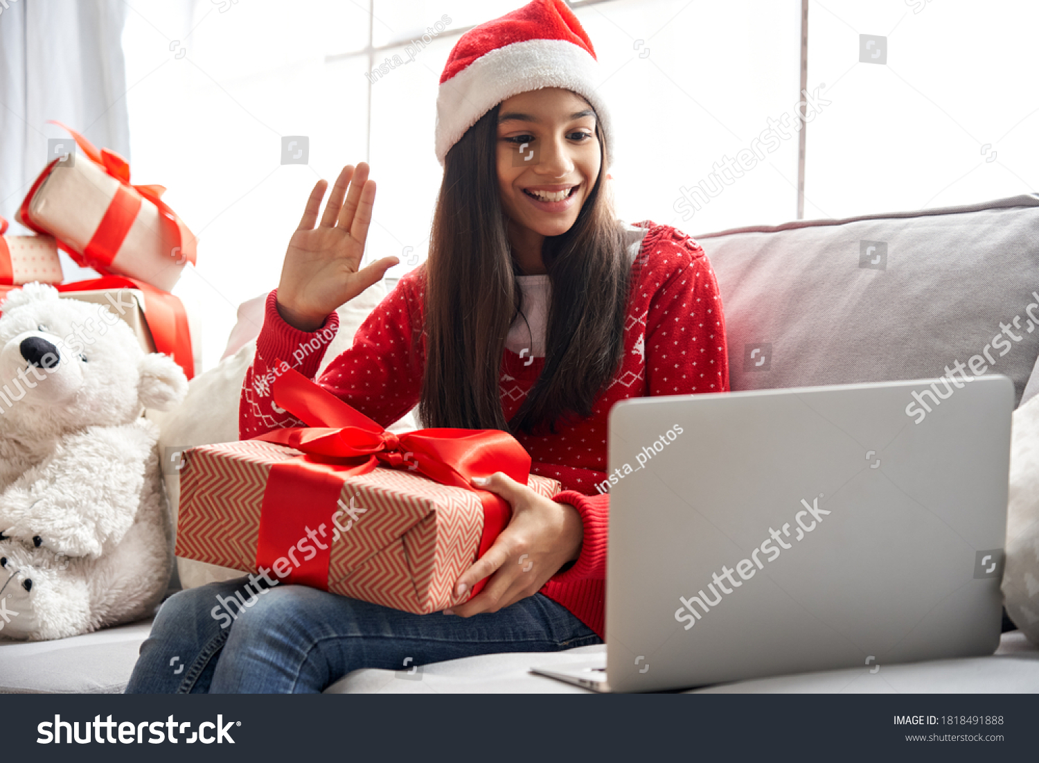 Smiling indian latin child kid girl wearing santa hat holding Christmas gift box, New Year present waving hand video calling family by webcam virtual meeting chat on holiday sitting on couch at home. #1818491888