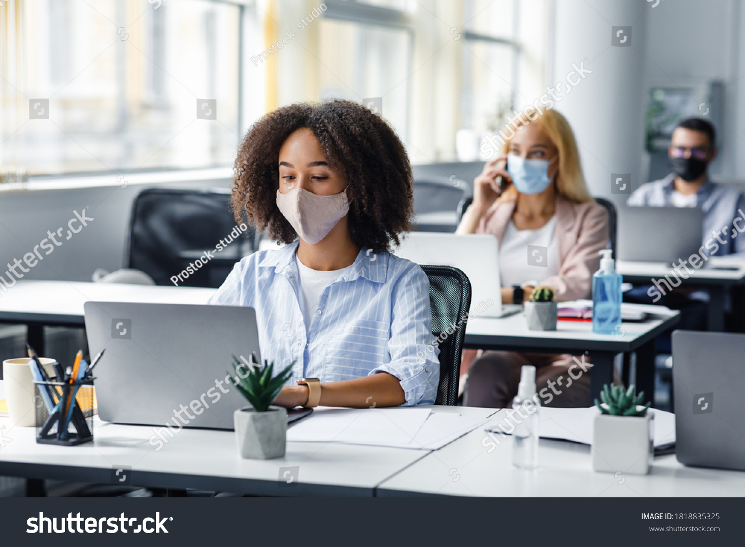 Social distancing at coronavirus outbreak situation. Pretty african american lady in protective mask typing on laptop, working for company support center in office interior with other employees #1818835325