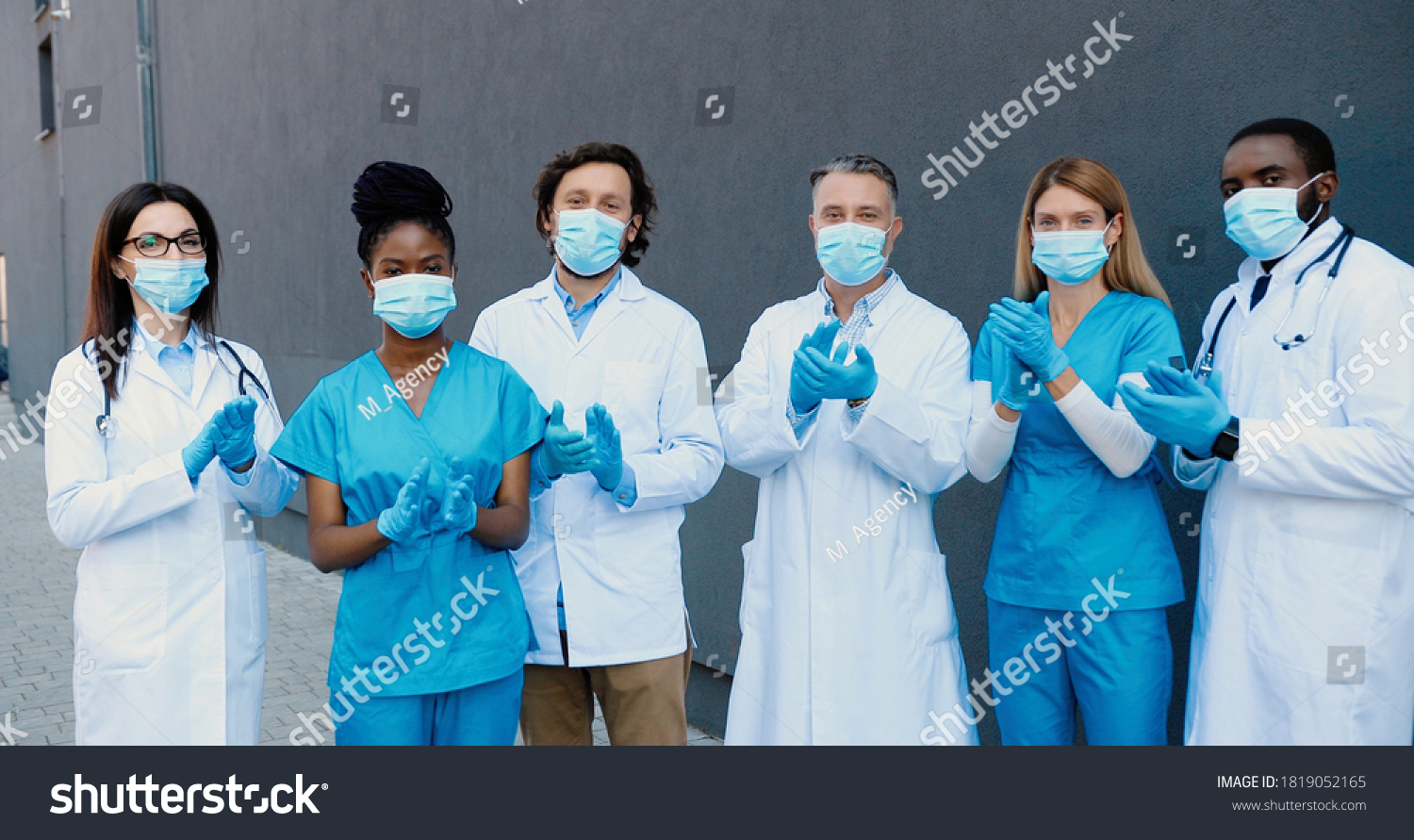Portrait of shot mixed-races young men and women doctors in medical masks, gloves and white gowns standing, applauding and looking at camera. Multi-ethnic medics. Covid-19 victory concept. Applause. #1819052165