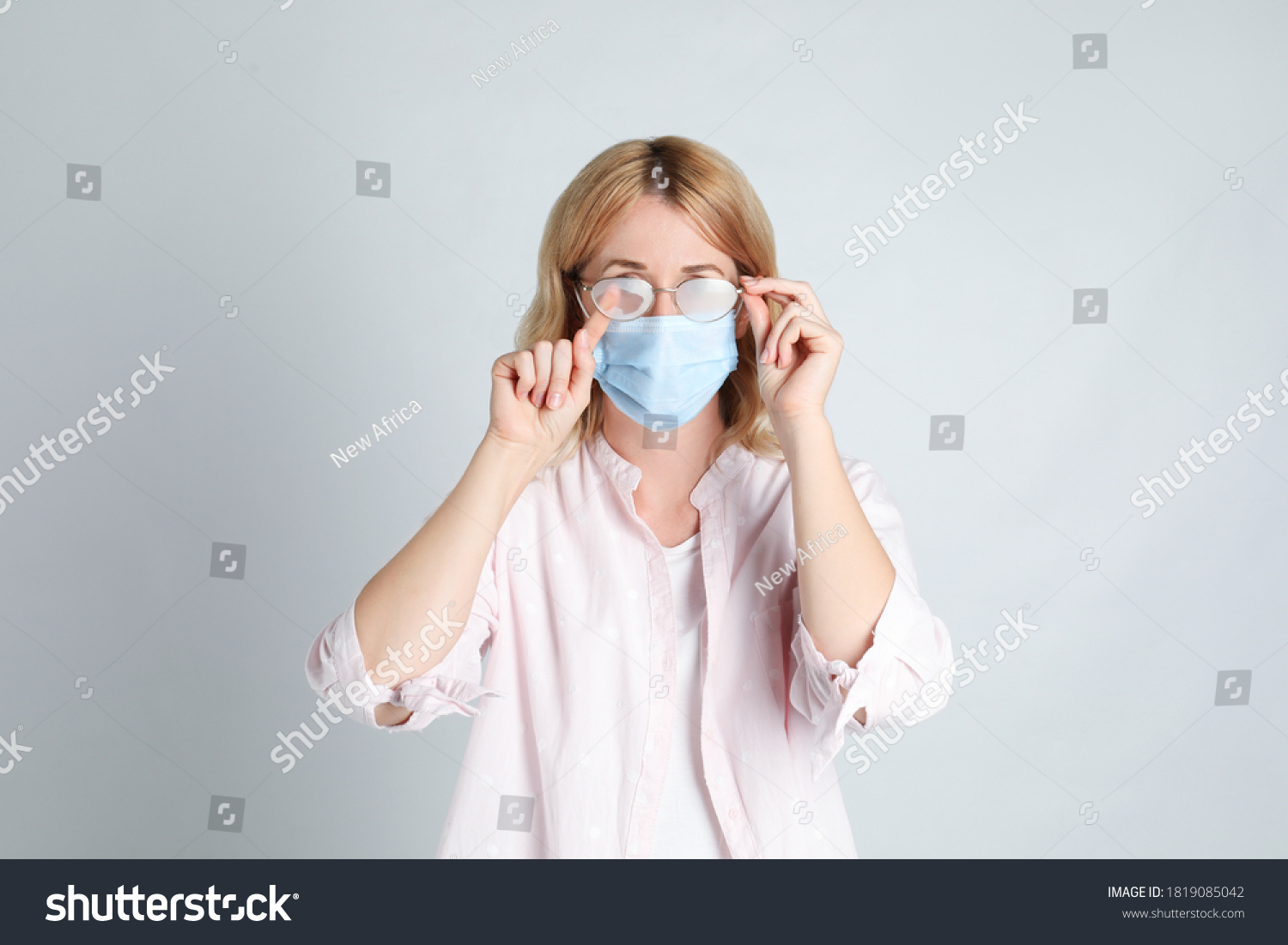 Woman wiping foggy glasses caused by wearing medical mask on light background #1819085042