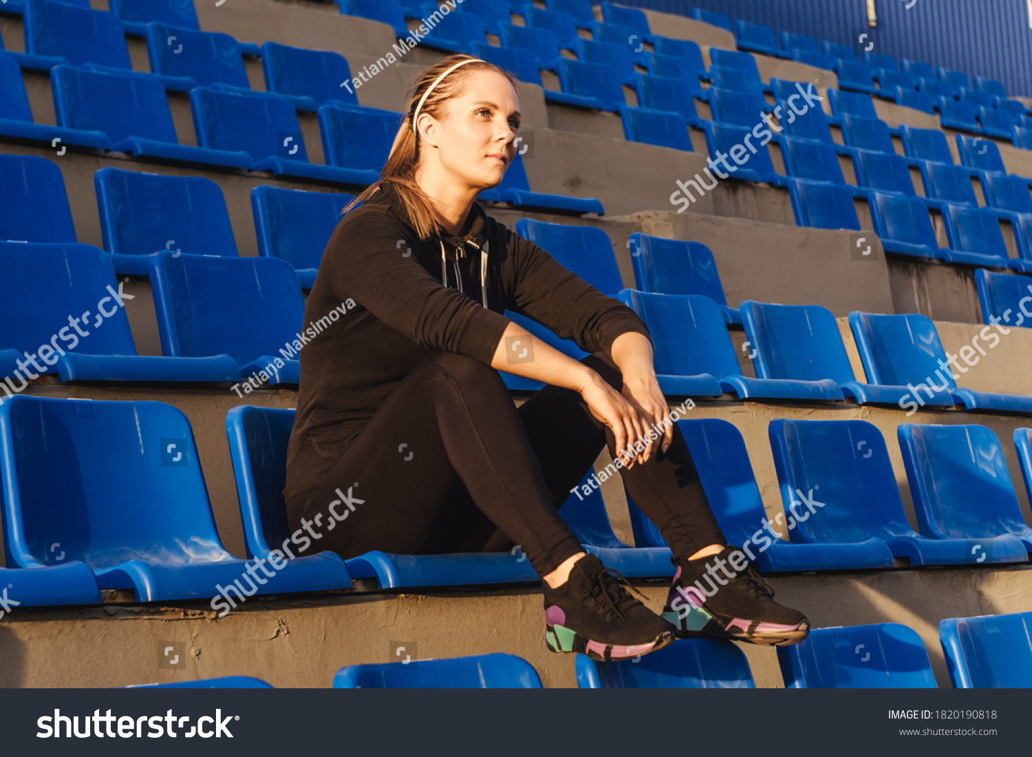 Fitness woman in black tracksuit sitting on the podium of the stadium. Woman after training. Sunset light, full length. #1820190818