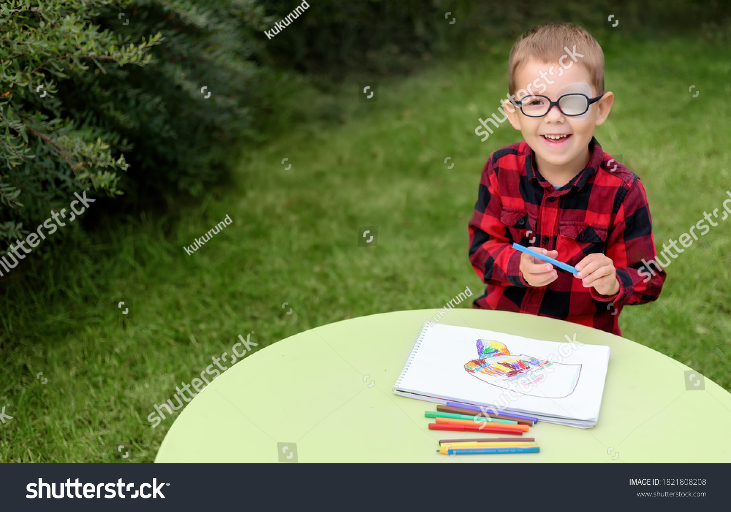 A little boy drawing a picture in the garden (outdoors). Wearing glasses and an eye patch (plaster, occluder) to prevent amblyopia and strabismus (squint, lazy eye). Child vision disease problem. #1821808208