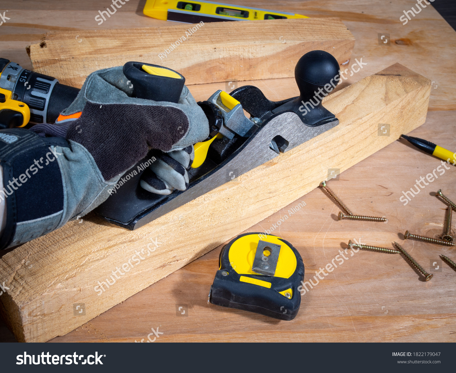 Hand in a safety glove keeping plane tool above the wooden bar, tape measure, screws and instruments around.  #1822179047