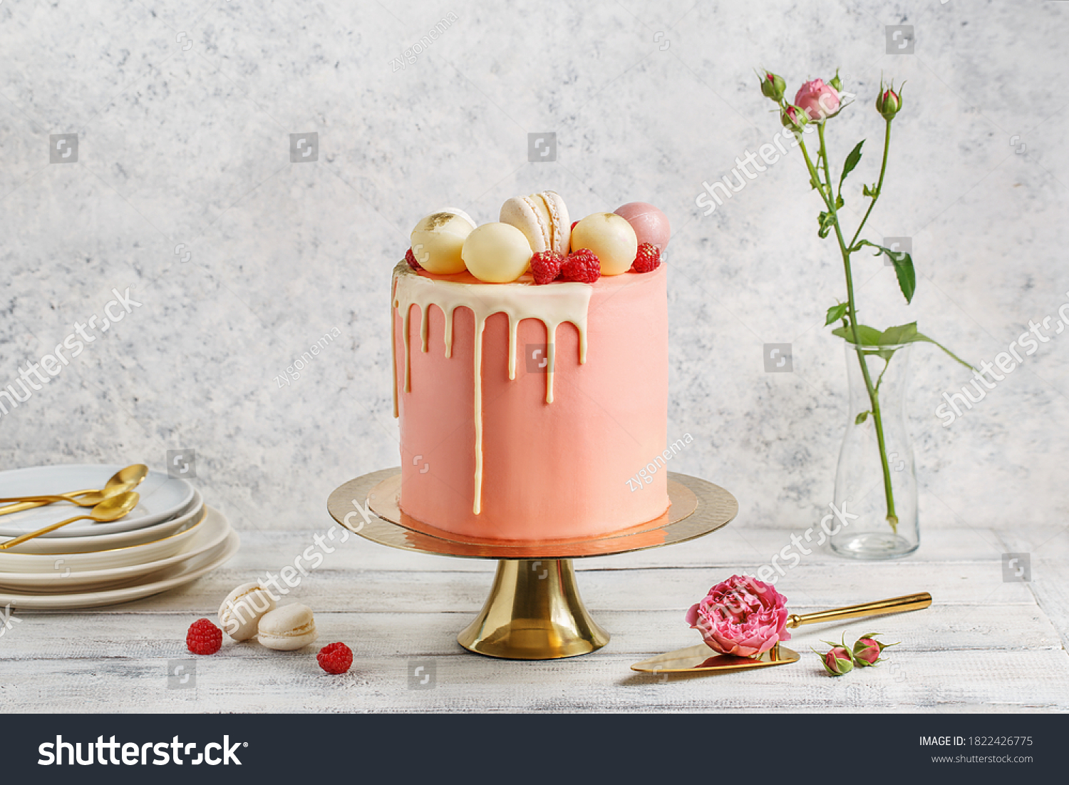 Tall pink cake decorated with macaroons, raspberries and chocolate balls on golden cake stand over white background with flowers and berries. Side view, copy space #1822426775