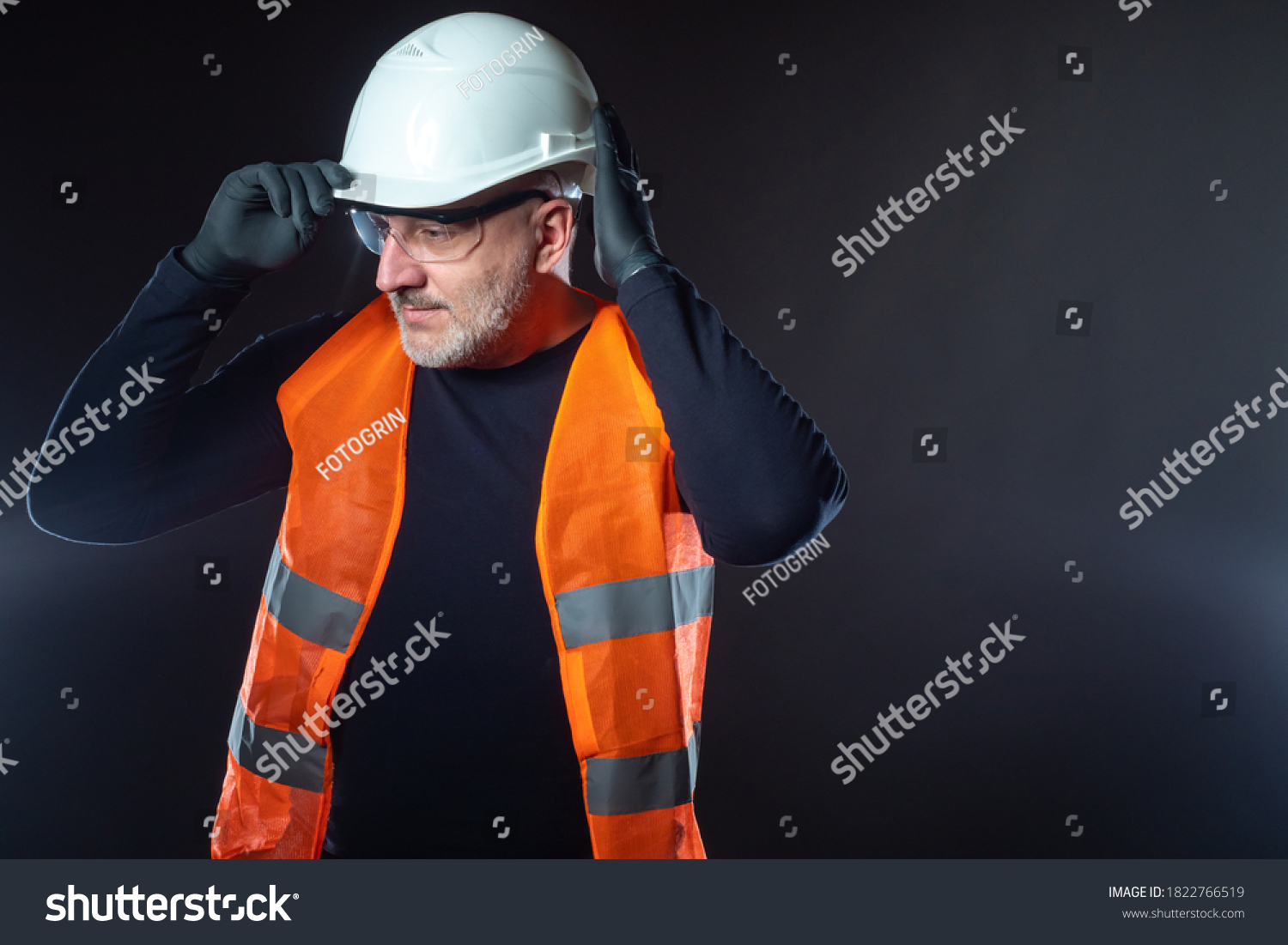 Portrait of the Builder on a dark background. A man in an orange vest puts a white construction helmet on his head. Construction uniforms. Jobs at the construction site. Place for text. #1822766519