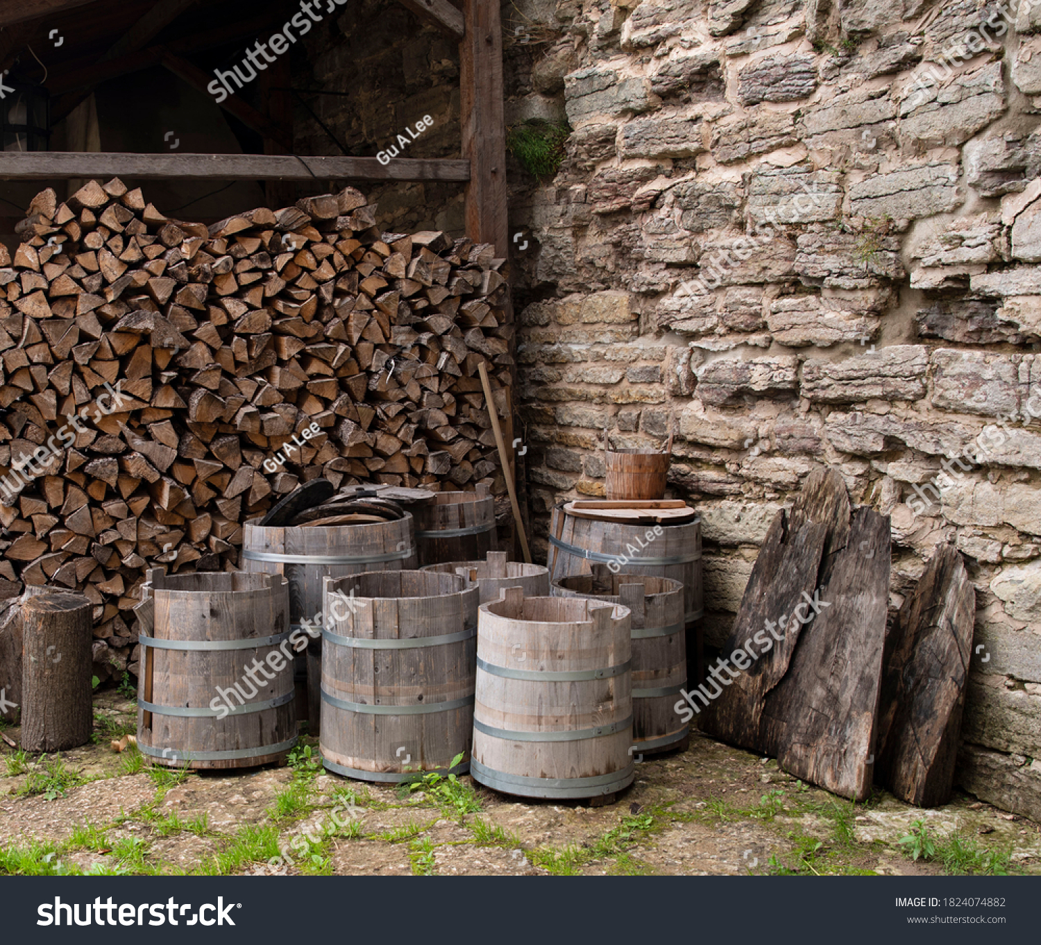There are barrels and tubs near the wall of the medieval castle, in the background there is a woodpile of chopped wood for the stove. #1824074882