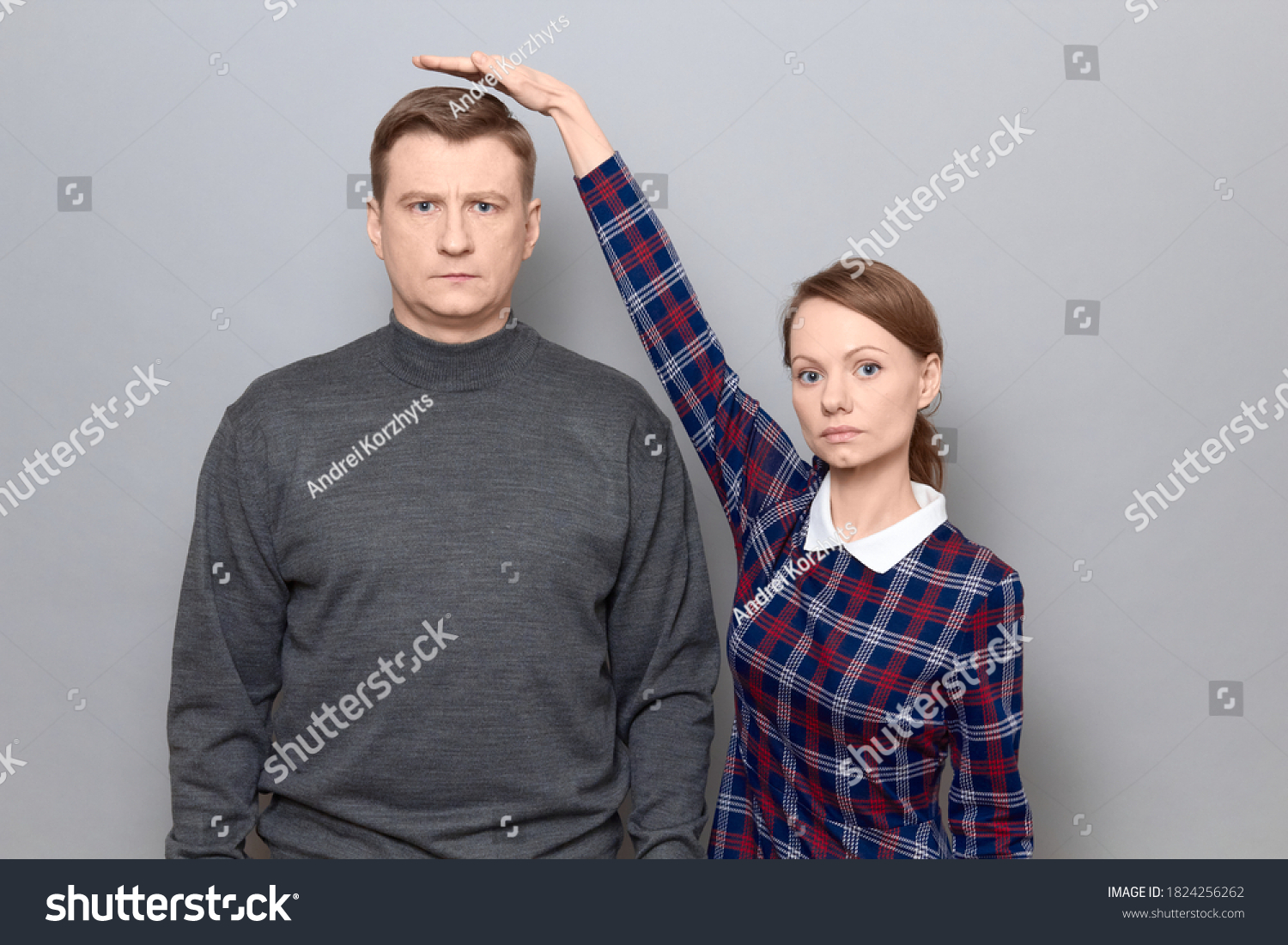 Studio shot of short woman standing on tiptoes and showing height of tall man, both are with serious expressions, over gray background. Concept of diversity of people's heights, tall and short persons #1824256262