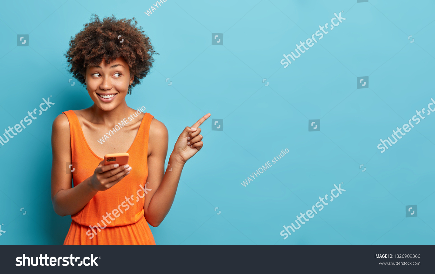 Nice attractive young Afro American woman uses cell phone and points on copy space shows direction dressed in stylish orange dress isolated over blue background. Ethnic lady with modern device #1826909366