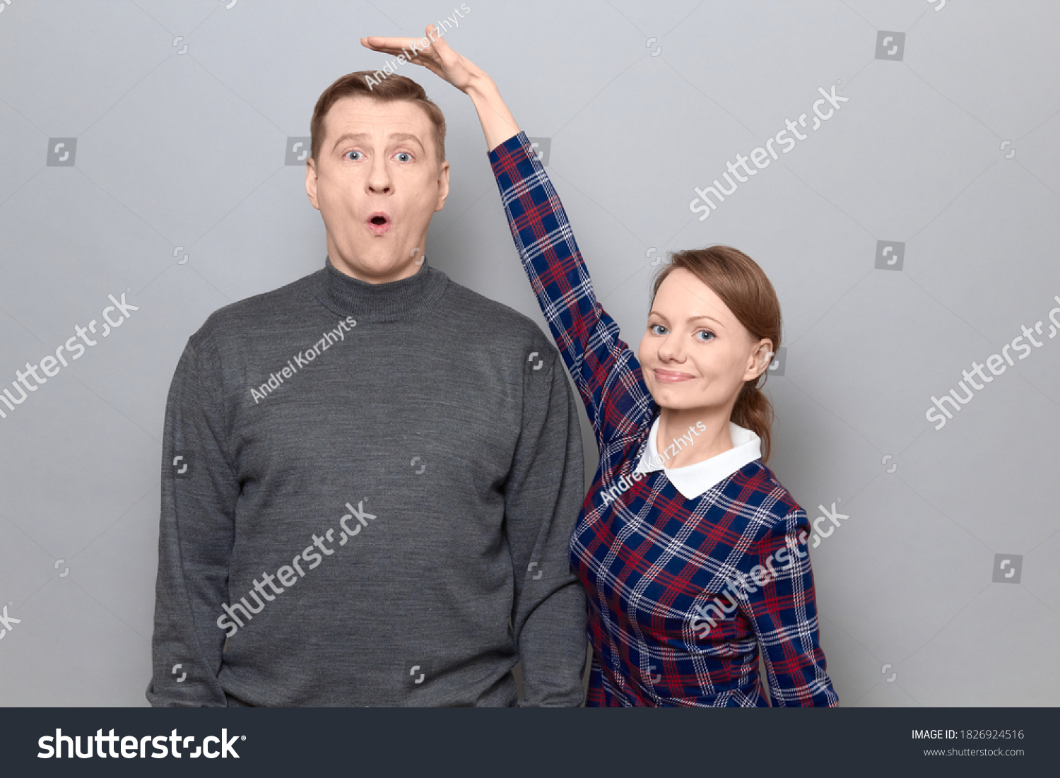 Studio shot of short woman standing and showing height of tall man, woman is smiling, man is amazed, over gray background. Concept of diversity of people's heights, tall and short persons #1826924516