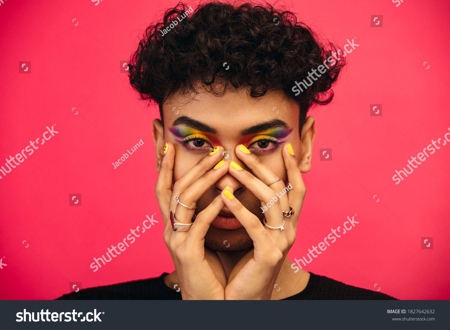 Gender fluid male wearing rainbow colored eye shadow and smiley face on fingernail. Transgender male with funky makeup on red background. #1827642632