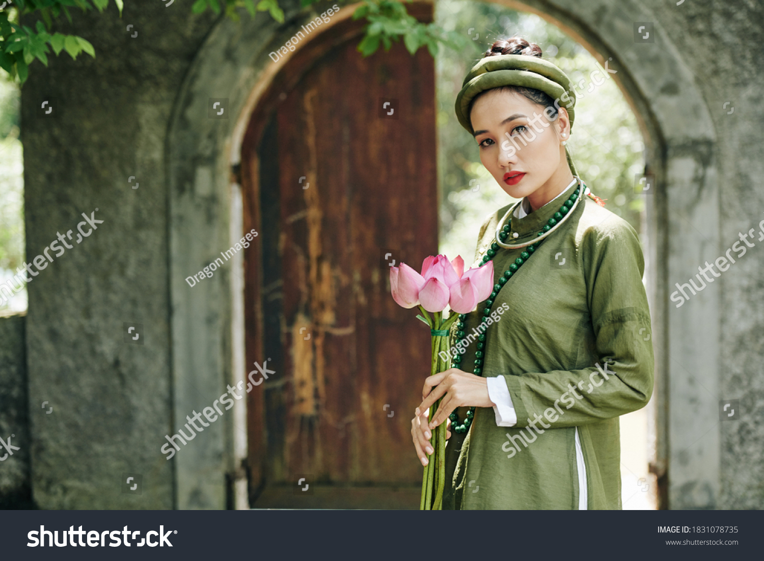 Portrait of attractive serious young woman in traditional Vietnamese dress and headwear holding lotus flowers and looking at camera #1831078735