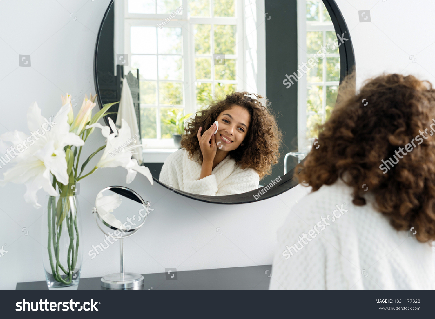 Natural beauty concept. Glad young afro american woman making morning routine, removing makeup, looking at mirror, holding cotton pad in hands near face and sitting in bathrobe at bathroom #1831177828