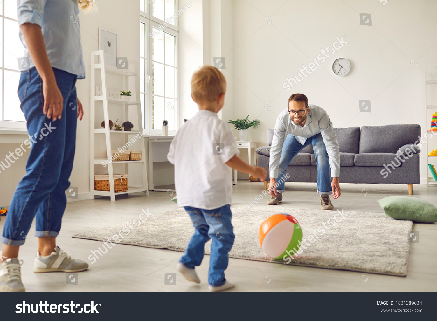Happy family staying at home, having fun together and engaging their small child in sport games. Young dad with little son playing football with inflatable ball in living-room #1831389634