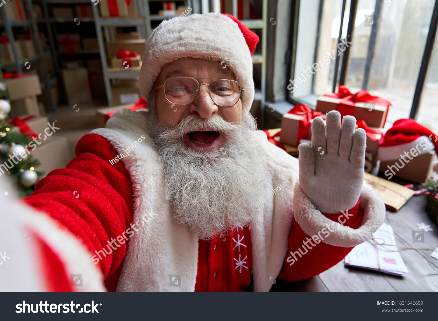 Happy old bearded Santa Claus wearing costume holding phone waving hand taking selfie, video calling, recording video Merry Christmas greeting or shooting vlog standing in workshop, face camera view. #1831546699