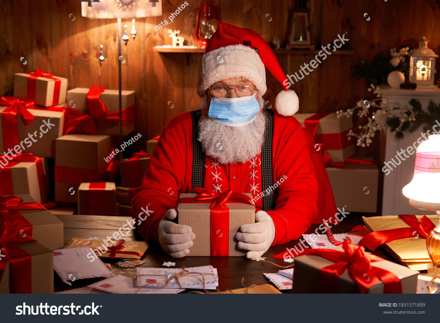 Old funny bearded Santa Claus wearing face mask, holding gift box preparing for xmas eve sitting at cozy home table late in night with presents. Merry Christmas Covid 19 coronavirus safe delivery. #1831571899