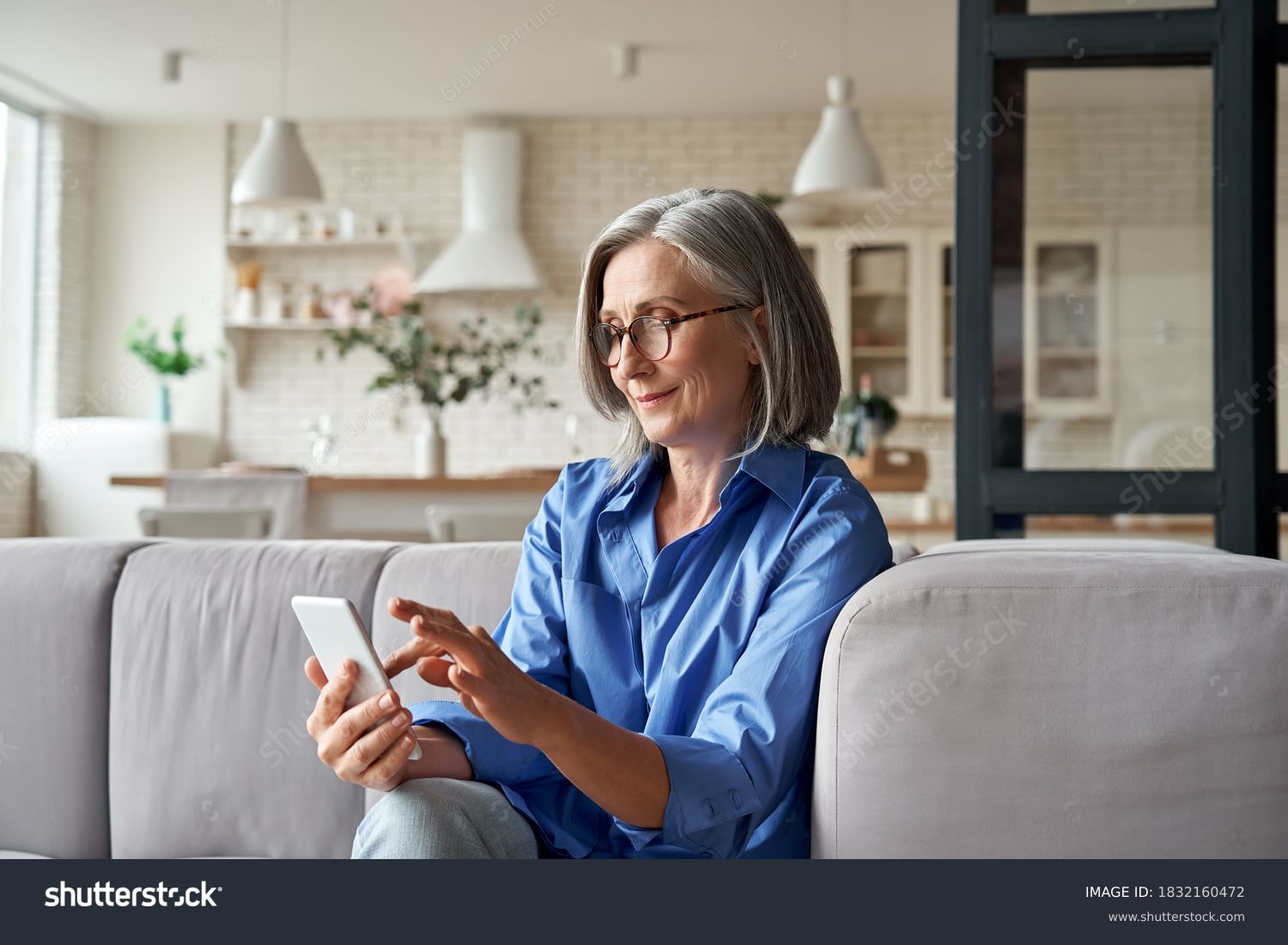 Relaxed mature old 60s woman, older middle aged female customer holding smartphone using mobile app, texting message, search ecommerce offers on cell phone technology device sitting on couch at home. #1832160472